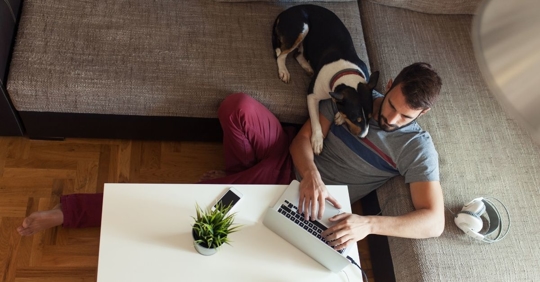 man sitting on the floor in front of his couch, typing on a computer, medium size dog is resting head on man's shoulder